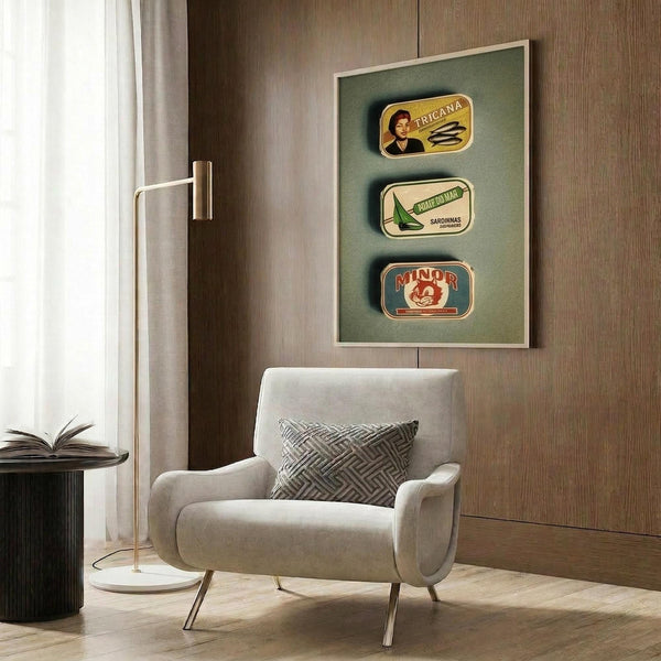 May include: Three vintage Portuguese sardine tin labels displayed in a light wooden frame against sage green background on wood-paneled wall. Modern cream armchair with geometric pillow sits below artwork next to brass floor lamp and dark round side table with open book. Natural lighting from window with white curtains.