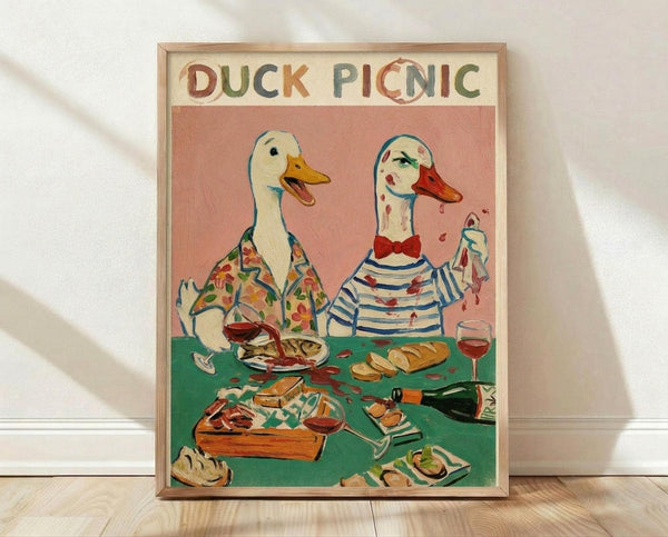 May include: Framed poster print leaning against white wall on light wood floor. Artwork shows two anthropomorphic ducks having a picnic at green table with wine glasses, bread, and food. Left duck wears floral shirt, right duck in striped shirt with red bow tie has disheveled appearance with red stains. Pink background with "DUCK PICNIC" text in colorful letters. Natural wood frame visible.