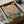 May include: Vintage-style poster print featuring two anthropomorphic ducks having a picnic, displayed in black frame inside cardboard shipping box with protective wrapping. Left duck wears floral shirt, right duck in striped top and red beret holds wine glass. Green picnic blanket shows bread, wine bottles, and food items. Pink background with "DUCK PICNIC" text at top. Whimsical retro illustration style with muted colors.