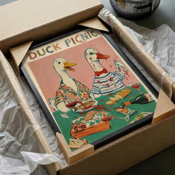 May include: Vintage-style poster print featuring two anthropomorphic ducks having a picnic, displayed in black frame inside cardboard shipping box with protective wrapping. Left duck wears floral shirt, right duck in striped top and red beret holds wine glass. Green picnic blanket shows bread, wine bottles, and food items. Pink background with "DUCK PICNIC" text at top. Whimsical retro illustration style with muted colors.