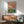 May include: Whimsical vintage-style poster featuring two anthropomorphic ducks in human clothing having a picnic, displayed in wooden frame above mid-century modern credenza with white ceramic lamp. Green upholstered armchair with wooden arms sits nearby in bright, minimalist room with white walls and natural lighting creating geometric shadows.