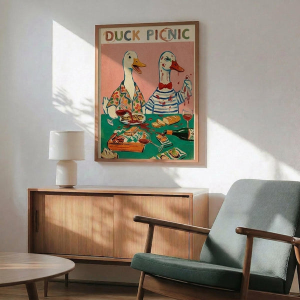 May include: Whimsical vintage-style poster featuring two anthropomorphic ducks in human clothing having a picnic, displayed in wooden frame above mid-century modern credenza with white ceramic lamp. Green upholstered armchair with wooden arms sits nearby in bright, minimalist room with white walls and natural lighting creating geometric shadows.