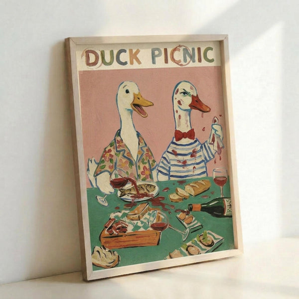 A framed art print leaning against a white wall showing two anthropomorphic ducks having a picnic. The left duck wears a floral shirt while the right duck wears a striped top with a red bow tie. Both have wine glasses and are seated at a green picnic blanket covered with food including bread, cheese, and other picnic items. The background is pink and the title "DUCK PICNIC" appears at the top in colorful letters. The artwork has a whimsical, illustrated style in a light wood frame.