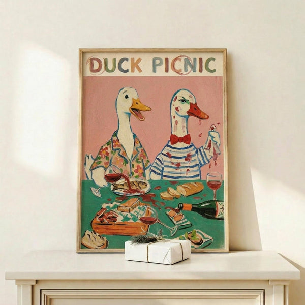 May include: Framed poster art showing two anthropomorphic ducks in human clothing at a picnic table. One duck wears a floral shirt, the other a striped top with bow tie. The table displays food, wine glasses, and a wine bottle on a green surface against a coral pink background. The colorful text "DUCK PICNIC" appears at the top. The wooden-framed artwork sits on a light wood shelf next to a small white wrapped gift.