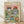 May include: Framed poster print leaning against white wall on light wood floor. Colorful illustrated artwork shows two cats wearing sunglasses and hats at outdoor picnic - one in fish-print shirt holding wine glass, other in striped shirt holding fish. Pink sky background with green grass foreground. Picnic spread includes wine bottle labeled "Catnip Party," plates of fish, bread basket, bowl of kibble, wine glasses, and cat toys scattered around.