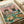 May include: Vintage-style poster print displayed on white paper showing a tabby cat in floral shirt and sunglasses alongside a French bulldog in striped shirt at a lunch table. Pink and green illustration features various foods including fish, bread, cheese, and wine bottle in retro art style.
