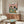 May include: Whimsical illustrated poster showing a tabby cat in floral shirt and French bulldog in striped shirt sitting at green table with fish, bone, cheese, bread and wine. Poster has cream background with pink section, displayed above walnut wood credenza in modern living room with white curved chair and glass coffee table.