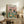 May include: Framed poster showing a tabby cat in sunglasses and floral shirt sitting with a French bulldog in striped shirt and bow tie at a dining table. The illustrated scene features fish and bone on plates, cheese, bread, wine, and appetizers spread across a teal tablecloth against a pink background. The wooden-framed artwork sits on a rustic wood sideboard alongside books, a ceramic vase, and fern plant in a cozy living room setting.