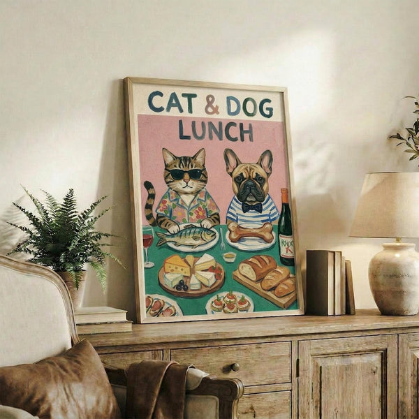 May include: Framed poster showing a tabby cat in sunglasses and floral shirt sitting with a French bulldog in striped shirt and bow tie at a dining table. The illustrated scene features fish and bone on plates, cheese, bread, wine, and appetizers spread across a teal tablecloth against a pink background. The wooden-framed artwork sits on a rustic wood sideboard alongside books, a ceramic vase, and fern plant in a cozy living room setting.