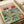 Two stylized cats wearing sunglasses enjoy a picnic on green grass under a yellow sun and pink sky. The left cat wears a straw hat and fish-print shirt while holding red wine, the right cat sports a red beret and blue striped shirt while holding a whole fish. The scene includes plates of fish, a bowl of cat food, bread and cheese basket, catnip party bottle, wine glasses, and colorful toys scattered around. The vintage-style illustration uses warm pastels and is printed on cream paper, photograp