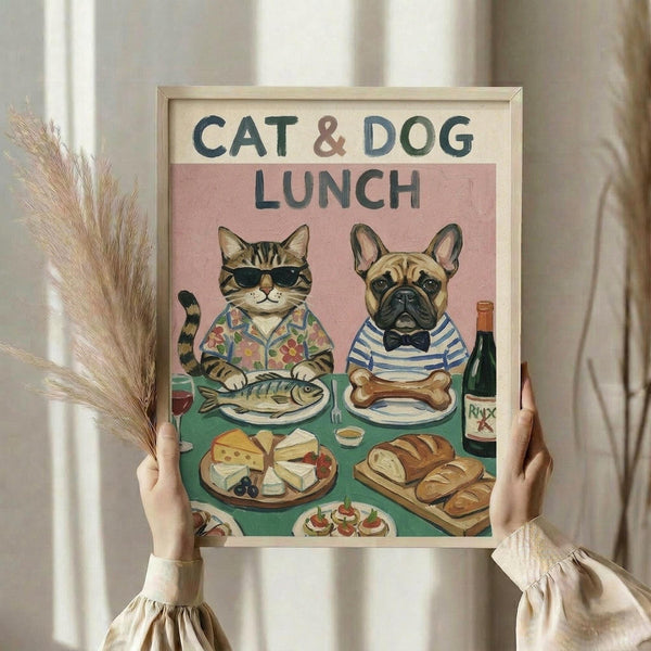 May include: Hands holding a framed poster featuring a tabby cat in sunglasses and floral shirt alongside a French bulldog in striped shirt and bow tie, both seated at a green table with fish, bone, cheese, bread and wine. Pink background with "CAT & DOG LUNCH" text. Light wood frame, dried wheat stems visible, neutral room setting with curtains.