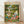 May include: Framed poster print leaning against white wall on light wooden floor. Artwork shows two white ducks wearing sunglasses in a park setting - one in floral shirt doing yoga pose on blue mat, another in striped shirt relaxing on pink blanket. Scene includes wine bottles, glasses, picnic basket, and colorful flowers scattered around green grass with trees in background. Light wood frame, vintage illustration style with warm colors.