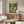 May include: A framed poster showing cartoon ducks in a park setting with colorful "QUACK & CHILL" text at top. One duck wears sunglasses and a floral shirt while another does yoga on a pink mat. The artwork features bright greens, blues, and warm tones. Displayed above a mid-century modern walnut credenza with white table lamp, next to a cream swivel chair and glass-top coffee table in a minimalist living room with white walls and natural lighting.