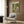 May include: Framed poster on wood-paneled wall showing cartoon ducks doing yoga in a park setting. One duck wears sunglasses and floral shorts while stretching, another lies on a yoga mat. Colorful illustration with green trees, picnic scene, and "QUACK & CHILL" text at top. Room features beige armchair with geometric pillow, brass floor lamp, dark side table with open book, white curtains, and wooden flooring.