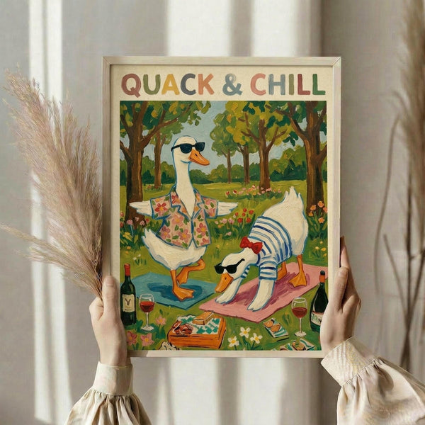 May include: Hands holding a framed art print featuring two white ducks wearing sunglasses and colorful clothing in a park setting with trees, flowers, wine glasses, and a picnic blanket. The poster has "QUACK & CHILL" text at the top in multicolored letters. Light wooden frame against white curtain background with dried pampas grass visible on the left side.