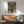 May include: Framed poster displaying anime-style illustration of person in brown jacket standing before large mech robot in cyberpunk cityscape with green and orange tones, featuring Japanese and Korean text at top. Shown hanging above mid-century modern wooden credenza with white ceramic lamp, next to teal upholstered chair with wooden arms in sunlit room with white walls.