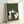 May include: A framed print leaning against a white wall showing six cats of various colors sitting around a green poker table playing cards. The artwork features a traditional Japanese woodblock print style with fine line work and muted colors against a sage green textured background. The light wooden frame complements the earthy tones of the illustration.