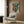 May include: Framed anime-style poster featuring a soldier in green military gear standing before a large brown and teal mech robot, displayed on wood paneled wall above modern cream armchair with geometric pillow, brass floor lamp and side table with open book nearby