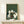 May include: A framed poster featuring five cats playing poker around a green table with playing cards scattered on top. The artwork has a Japanese woodblock print style with a dark green textured background. The cats include white, black, and striped varieties sitting on wooden chairs. The print is displayed in a light wood frame on a white shelf next to a small wrapped gift box against a cream colored wall.