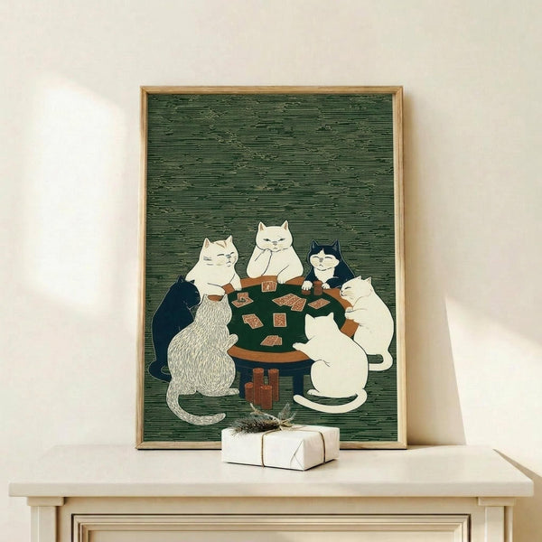 May include: A framed poster featuring five cats playing poker around a green table with playing cards scattered on top. The artwork has a Japanese woodblock print style with a dark green textured background. The cats include white, black, and striped varieties sitting on wooden chairs. The print is displayed in a light wood frame on a white shelf next to a small wrapped gift box against a cream colored wall.