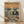 May include: Framed vintage-style poster print leaning against white wall on light wood floor. Features two koalas in sunglasses dining at seaside restaurant - one in tropical shirt and hat, other in black tuxedo. Eucalyptus wine bottle, fish dinner, cheese board visible on wooden table with ocean sunset backdrop. Warm orange and teal color palette in weathered illustration style.
