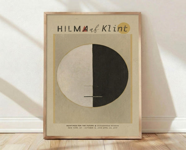 May include: Framed poster print leaning against white wall on light wood floor. Features beige background with circular design split between cream and black halves, small horizontal lines through center. Text reads "HILMA af Klint" at top with small red triangle accent, exhibition details at bottom. Natural wood frame, soft lighting creates diagonal shadows across scene.
