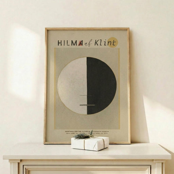 May include: Framed museum exhibition poster featuring Hilma af Klint's abstract geometric artwork - a circle split into white and black halves with thin horizontal lines, displayed on light wooden shelf with small wrapped gift box beside it against cream wall