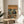 May include: Framed poster showing two koalas in sunglasses dining at a wooden table with wine and fish, set against an orange sunset beach scene. The left koala wears a tropical shirt and straw hat, the right wears a black tuxedo. Text reads "KOALA FINE DINING" at top. Frame sits on round wooden side table next to beige upholstered chair with plant shadows visible on white wall.