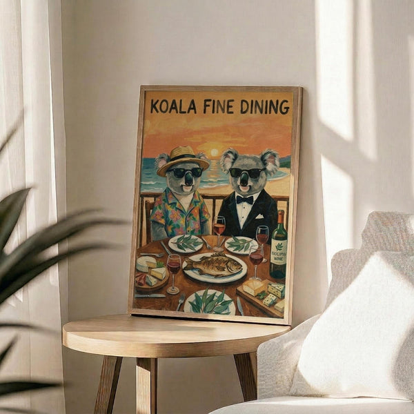 May include: Framed poster showing two koalas in sunglasses dining at a wooden table with wine and fish, set against an orange sunset beach scene. The left koala wears a tropical shirt and straw hat, the right wears a black tuxedo. Text reads "KOALA FINE DINING" at top. Frame sits on round wooden side table next to beige upholstered chair with plant shadows visible on white wall.