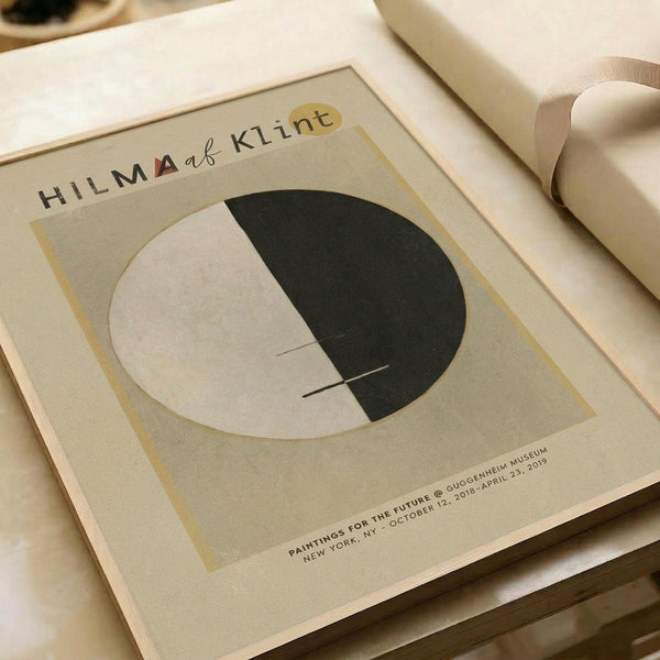 May include: Minimalist abstract art poster with beige background featuring a circular design split between cream and dark green halves with thin intersecting lines. The poster displays "HILMA af KLINT" text at top and Guggenheim Museum exhibition details at bottom. Shown in white matting alongside rolled paper tubes on a neutral surface.