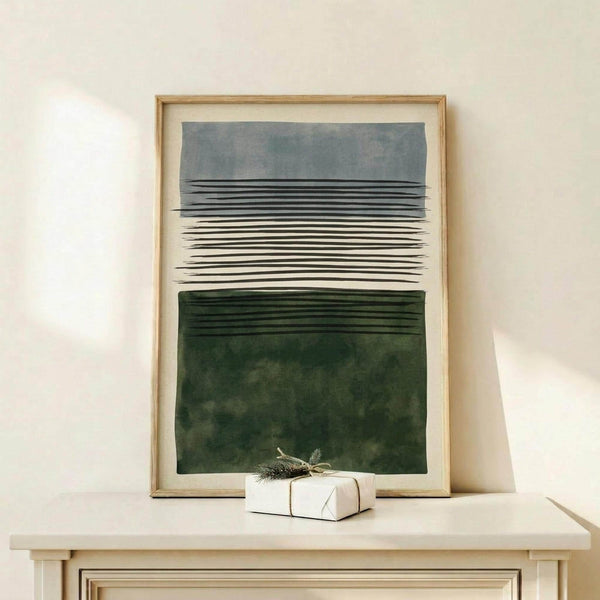 May include: Framed abstract art print with horizontal black lines transitioning from gray-blue at top to deep forest green at bottom, displayed on light wood console table against cream wall with small wrapped gift box decorated with greenery beside frame