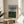 May include: A framed abstract art print leaning against a beige wall on a round wooden side table. The artwork features horizontal bands in blue-gray, cream, and forest green with black linear details creating a layered landscape effect. Natural wood frame with white matting. Soft natural lighting creates shadows across the neutral-toned room setting with a beige upholstered chair and plant leaves visible at the edges.