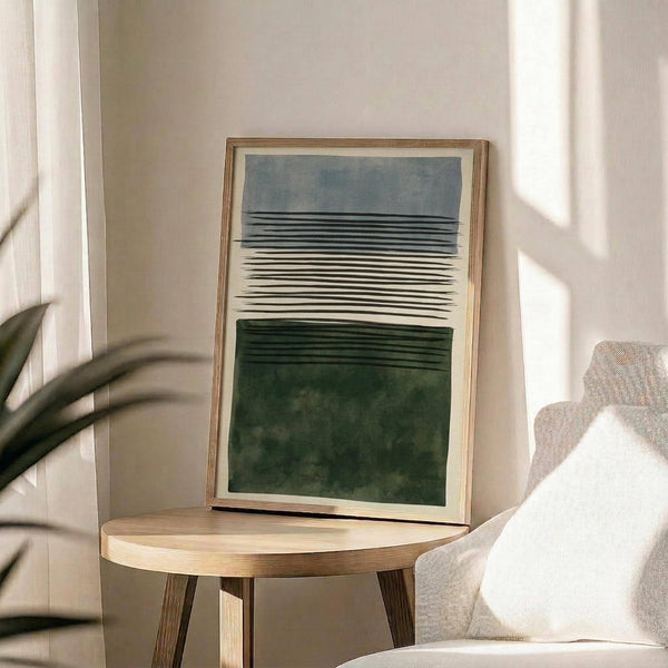 May include: A framed abstract art print leaning against a beige wall on a round wooden side table. The artwork features horizontal bands in blue-gray, cream, and forest green with black linear details creating a layered landscape effect. Natural wood frame with white matting. Soft natural lighting creates shadows across the neutral-toned room setting with a beige upholstered chair and plant leaves visible at the edges.