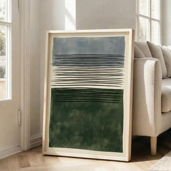 May include: Large framed abstract art print leaning against white wall next to beige upholstered chair. The artwork features horizontal black lines across the center with a gray watercolor wash above and deep green watercolor section below, creating a minimalist landscape effect. Natural wood frame with white matting, positioned on light wooden floors in bright, airy room with tall windows.