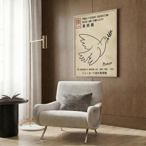 May include: Framed vintage-style cream poster featuring Picasso's simple line drawing of a dove carrying an olive branch, with exhibition text in English and Japanese characters. Poster hangs on wood-paneled wall above a beige mid-century armchair with geometric patterned pillow. Scene includes gold floor lamp and dark round side table with open book.