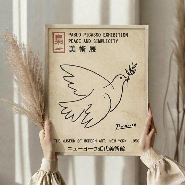 May include: Hands holding a framed beige poster featuring Picasso's minimalist line drawing of a dove carrying an olive branch, with exhibition text in English and Japanese. The scene shows dried pampas grass on the left and soft natural lighting against white curtains in the background.