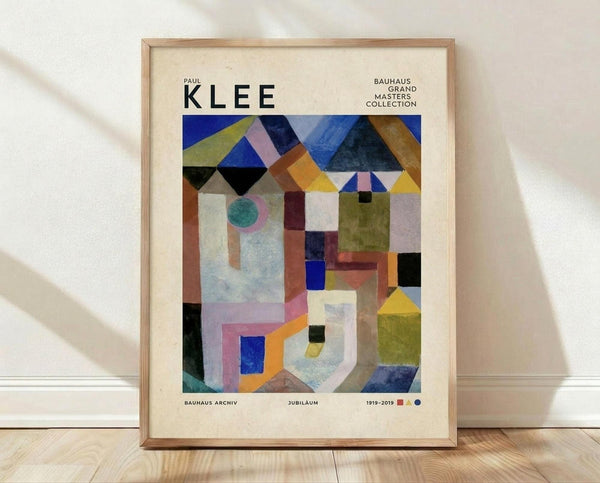 May include: A framed poster displaying Paul Klee's abstract geometric artwork with colorful triangular roofs, rectangular buildings, and shapes in blues, yellows, greens, and earth tones. The cream-colored print features "KLEE" at the top and "BAUHAUS ARCHIV JUBILÄUM 1919-2019" at the bottom. The natural wood frame leans against a white wall on light wooden flooring with soft shadows visible.