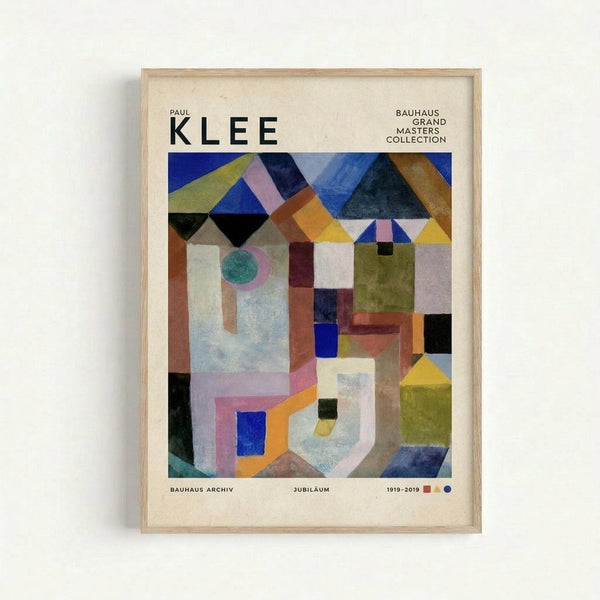 Framed abstract geometric art poster featuring Paul Klee's colorful composition with overlapping triangular shapes in blues, yellows, oranges, and greens at top, and angular architectural forms with circles and rectangles below. The artwork combines watercolor-like textures with bold geometric patterns. Text reads "PAUL KLEE" at top and "BAUHAUS GRAND MASTERS COLLECTION" in upper right, with "BAUHAUS ARCHIV JUBILÄUM 1919-2019" at bottom. Light wood frame against white wall.