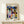 May include: Framed Paul Klee abstract geometric poster in light wood frame, featuring colorful overlapping shapes in blues, yellows, greens, and oranges. Print displays "KLEE" at top with "Bauhaus Grand Masters Collection" text. Artwork shows architectural forms, circles, triangles, and rectangles in Klee's distinctive style. Frame leans against white wall on light wooden surface next to small white wrapped gift with evergreen sprig.