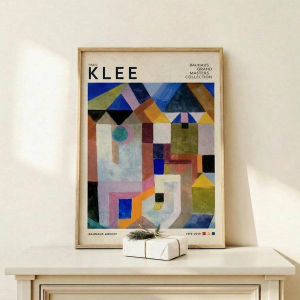 May include: Framed Paul Klee abstract geometric poster in light wood frame, featuring colorful overlapping shapes in blues, yellows, greens, and oranges. Print displays "KLEE" at top with "Bauhaus Grand Masters Collection" text. Artwork shows architectural forms, circles, triangles, and rectangles in Klee's distinctive style. Frame leans against white wall on light wooden surface next to small white wrapped gift with evergreen sprig.