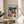 May include: Framed Paul Klee abstract art poster in light wood frame, displayed on round wooden side table next to beige upholstered chair. The colorful geometric artwork features overlapping shapes in blues, yellows, greens, and oranges with Bauhaus exhibition text. Soft natural lighting creates shadows across white walls, with partial view of plant leaves in corner.