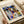 May include: Hands holding a framed art print featuring Paul Klee's abstract geometric composition with colorful overlapping shapes in blues, greens, oranges, and purples. The poster shows "PAUL KLEE" text at top and "BAUHAUS ARCHIV" at bottom, displayed on a neutral workspace with rolled paper in background. The artwork features triangular and rectangular forms creating a complex layered pattern.