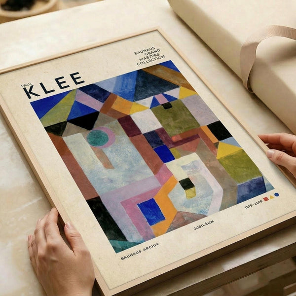 May include: Hands holding a framed art print featuring Paul Klee's abstract geometric composition with colorful overlapping shapes in blues, greens, oranges, and purples. The poster shows "PAUL KLEE" text at top and "BAUHAUS ARCHIV" at bottom, displayed on a neutral workspace with rolled paper in background. The artwork features triangular and rectangular forms creating a complex layered pattern.