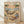 May include: Framed poster print leaning against white wall on light wood floor. Artwork shows two cats relaxing on tropical beach - one tabby in Hawaiian shirt sleeping in hammock between palm trees with "Z" sleep symbols, another orange tabby in striped shirt lounging on beach chair wearing sunglasses. Beach scene includes martini glass, fish skeleton, small sign reading "THE ART OF DOING NOTHING." Sunset sky with pink and orange hues over ocean. Vintage illustration style in warm tone