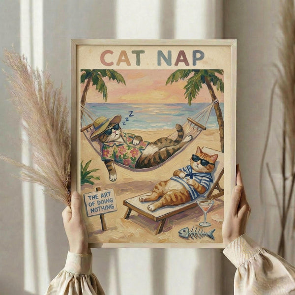 May include: Hands holding framed vintage-style poster showing two cartoon cats relaxing on tropical beach. One orange tabby sleeps in hammock wearing Hawaiian shirt and sun hat, another lounges on beach chair with sunglasses and striped shirt. Scene features palm trees, ocean sunset, cocktail glass, fish skeleton, and small sign reading "The Art of Doing Nothing." Light wood frame, pampas grass visible on left, soft neutral curtains in background.