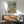 May include: Framed poster showing two ducks doing yoga on a beach scene with colorful text "Duck Yoga" at top. One duck in floral yoga outfit doing tree pose, another in striped shirt holding wine glass. Beach background with palm tree, ocean waves, yoga mat, wine bottles and snacks. Displayed above mid-century modern wooden credenza with white ceramic lamp, next to teal upholstered armchair in bright living room with natural sunlight.