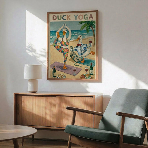 May include: Framed poster showing two ducks doing yoga on a beach scene with colorful text "Duck Yoga" at top. One duck in floral yoga outfit doing tree pose, another in striped shirt holding wine glass. Beach background with palm tree, ocean waves, yoga mat, wine bottles and snacks. Displayed above mid-century modern wooden credenza with white ceramic lamp, next to teal upholstered armchair in bright living room with natural sunlight.