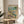 May include: Framed poster showing cartoon ducks doing yoga poses on a beach scene with palm tree, wine bottles and snacks scattered around. One duck in colorful patterned leggings holds tree pose on purple yoga mat while another duck in striped shirt relaxes with wine glass. Wooden frame sits on round wooden side table next to beige upholstered chair with decorative pillow. Soft natural lighting and plant leaves visible.