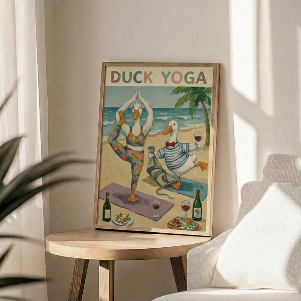 May include: Framed poster showing cartoon ducks doing yoga poses on a beach scene with palm tree, wine bottles and snacks scattered around. One duck in colorful patterned leggings holds tree pose on purple yoga mat while another duck in striped shirt relaxes with wine glass. Wooden frame sits on round wooden side table next to beige upholstered chair with decorative pillow. Soft natural lighting and plant leaves visible.