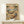 May include: Framed poster showing two cartoon cats relaxing on a tropical beach - one sleeping in a hammock wearing Hawaiian shirt and hat with "Z" sleep bubble, another lounging on beach chair with sunglasses and striped shirt holding cocktail. Palm trees frame ocean sunset scene. Small sign reads "The Art of Doing Nothing." White gift box with greenery sits on light wood shelf below cream-matted artwork in natural wood frame against white wall.